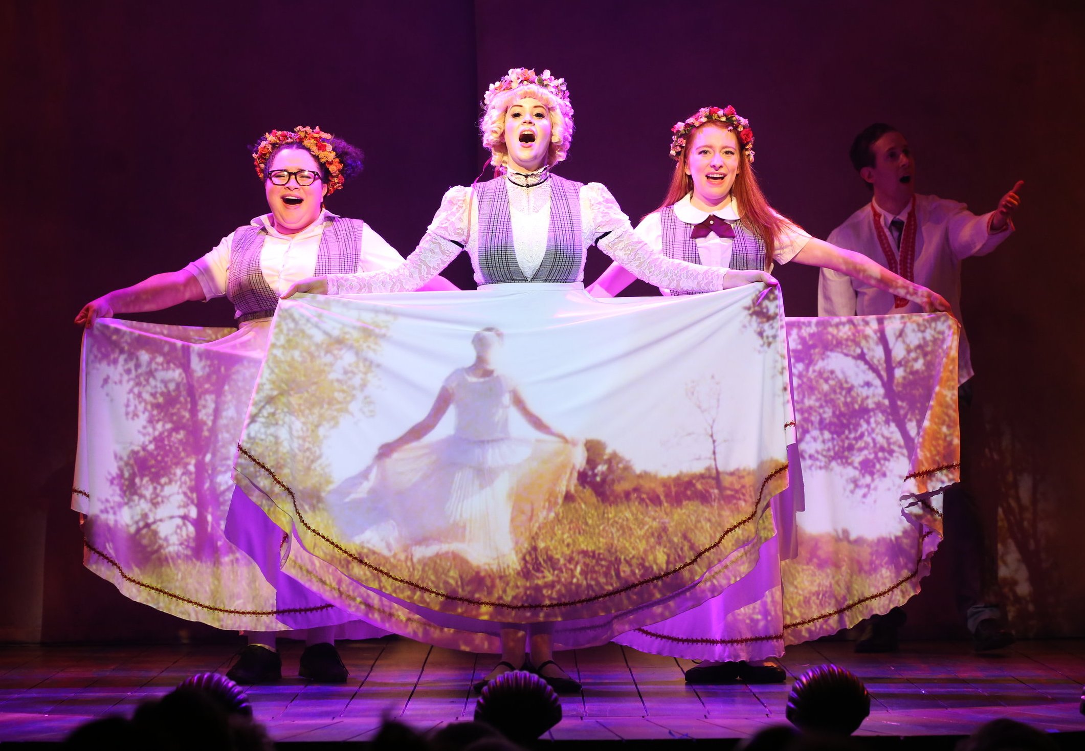 Constance, Jane, and Ocean dancing. They are wearing longer skirts, as well as flower crowns. They hold their long skirts as they dance, as a video of Talia in a field is projected onto them.